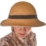 Front-facing view of a person wearing a straw-hat-style bicycle helmet with a black ribbon band. The broad brim slightly shades their eyes, and pastel pink curls frame the sides of their face. They wear a light-colored blouse printed with owl illustrations.