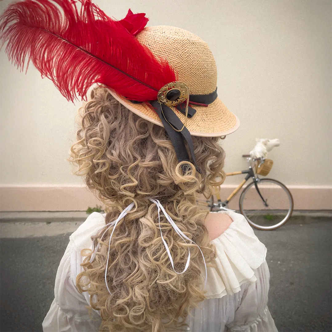 Back view of the Hair Helmet 1789, featuring a curly wig-style cover and a straw hat decorated with a large red feather, black ribbon, and gold brooch
