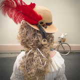Back view of the Hair Helmet 1789, featuring a curly wig-style cover and a straw hat decorated with a large red feather, black ribbon, and gold brooch