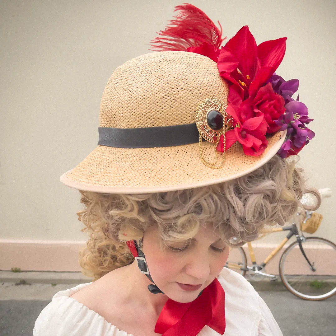 Woman wearing the Hair Helmet 1789, a bicycle helmet disguised with curly powdered wig-style hair and a vintage straw hat adorned with red flowers, purple blossoms, and a gold brooch