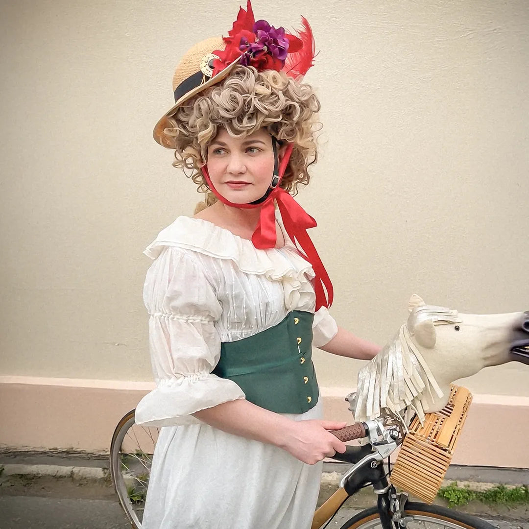 Woman dressed in 18th-century-inspired fashion wearing the Hair Helmet 1789, a bicycle helmet disguised with a curly wig and feathered straw hat, holding a vintage-style bike with a white horse head decoration