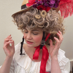 Woman adjusting the Hair Helmet 1789, a bicycle helmet styled with a curly powdered wig and feathered straw hat, blending 18th-century fashion with modern safety