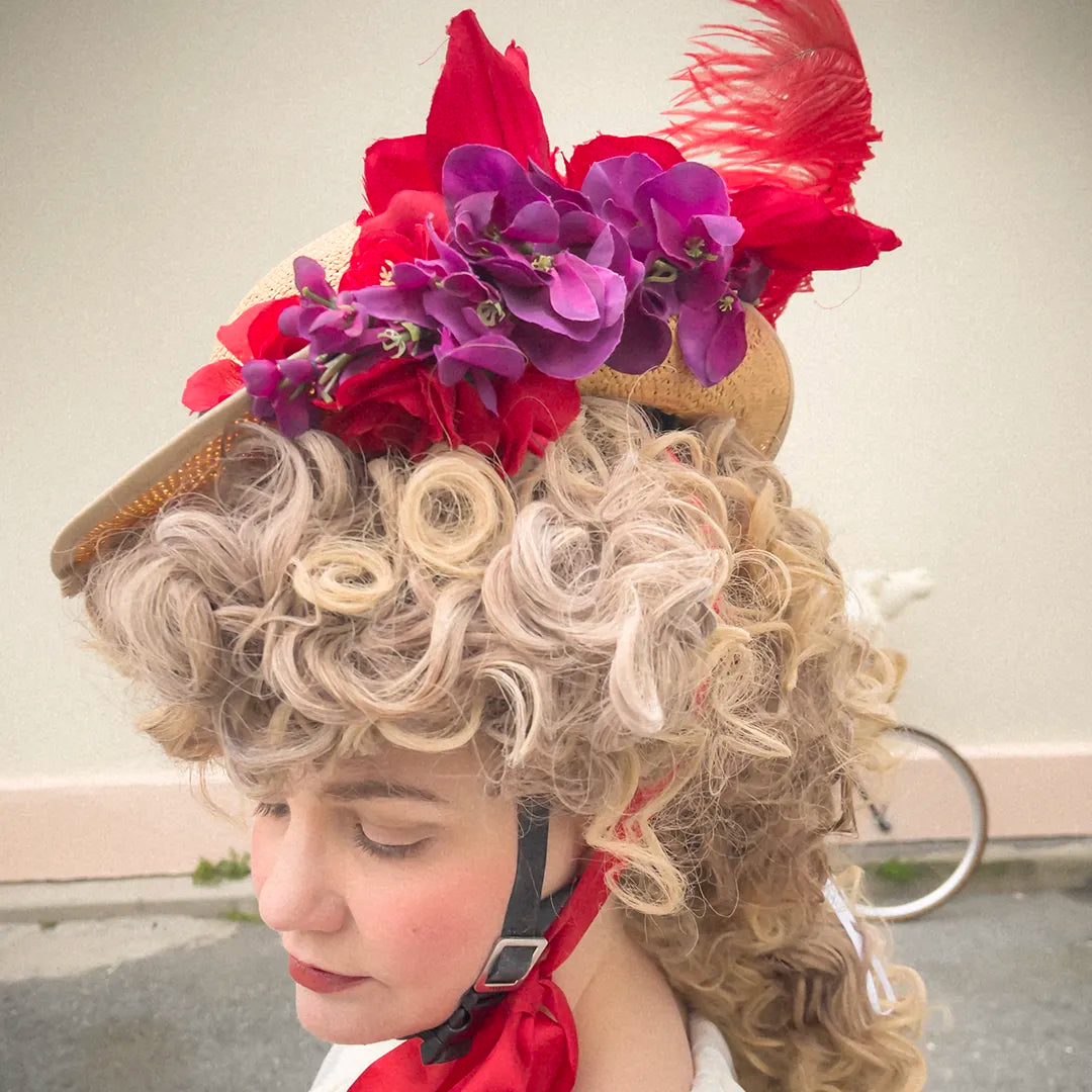 Side view of a woman wearing the Hair Helmet 1789, a bicycle helmet disguised with a voluminous curly wig and topped with red flowers, purple blossoms, and a red feather