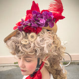 Side view of a woman wearing the Hair Helmet 1789, a bicycle helmet disguised with a voluminous curly wig and topped with red flowers, purple blossoms, and a red feather