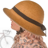 Side-profile view of a person wearing a straw-hat-style bicycle helmet with a black ribbon band. The woven texture of the helmet mimics a classic sun hat. The model has pastel pink hair and a patterned blouse, with a softly blurred bicycle silhouette in the background.