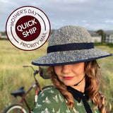 Melissa wearing a straw hat bike helmet with a navy band, smiling at the camera with her bike in the background. A circular badge overlay reads 'Mother's Day Quick Ship Priority Mail.'