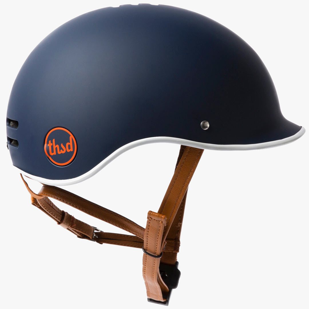 Vintage moto-inspired bicycle helmet in Navy blue with white and orange trim components. Viewed from the side on a plain white background. The tan chinstraps are visible below the rim of the helmet.